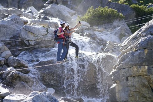 Two people cross a stream using a rope bridge, secured with a via ferrata set