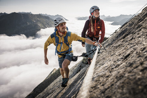 Zwei Klettersteiggeher überwinden eine einfache Passage am Klettersteig. Hinter ihnen liegt eine dicke Wolckendecke.