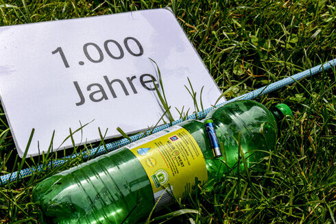 Eine Plastikflasche, eine Batterie und ein Schild mit der Aufschrift 1000 Jahre, liegen im Gras.