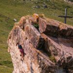 Das bild zeigt eine Klettererin am Hüttenblock bei der HEidelberger Hütte. Oben ein Holzkreuz, im Hintergrund grüne Wiesen.