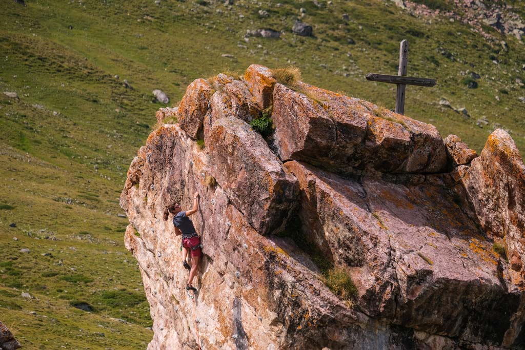 Das bild zeigt eine Klettererin am Hüttenblock bei der HEidelberger Hütte. Oben ein Holzkreuz, im Hintergrund grüne Wiesen.