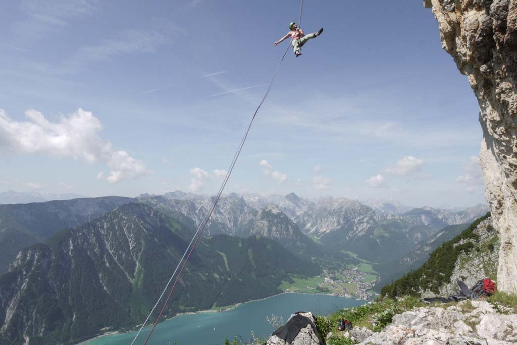 Das Bild zeigt einen Kletterer beim Abseilen über eine Felswand. Im Hintergrund der Achensee mit dem Dorf Achenkirch.